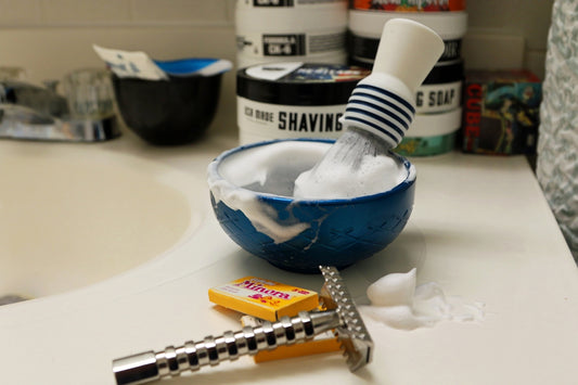 Shaving brush and bowl with lather and razor on sink.